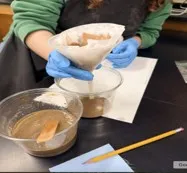 A photo showing a student wearing a green long-sleeve shirt, a black apron, and blue disposable gloves conducting a water-filtration activity at a lab table. They are holding a clear plastic funnel lined with layered filter papers, including pieces of brown paper, positioned above an empty clear plastic cup to catch the filtered liquid. In front of the student are two additional clear cups: One contains murky brown water with a wooden stir stick, and the other cup beneath the funnel is beginning to collect lighter, partially filtered water. A yellow pencil and a small blue cloth rest on the table nearby. 