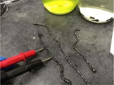 The photograph depicts worm-like, dark gray strings on a table, with multimeter prongs positioned to measure the resistance between prong ends.