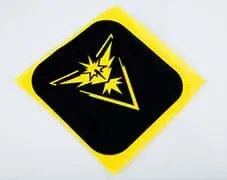 A photograph shows a square black iron-on fabric patch lying on a piece of yellow felt. Cut-outs in the patch let the yellow felt show through, revealing a Pokémon Zapdos species graphic that looks like a stylized bird with spread wings and spiky tail and head feathers.
