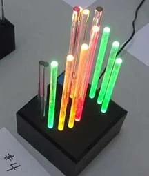 A photograph shows a student-created light sculpture composed of a dozen glowing clear sticks of different heights and colors positioned vertically in a black rack.