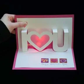 A photograph shows a hand opening a red and white pop-up and light-up valentine card that says “I <heart> you.”