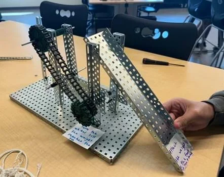 A photo showing a silver steel machine built with VEX parts that has a combination of wheels, gears, levers, and inclined plane. Printed signs are placed at two locations that have dark print on white background to give instructions to end users.