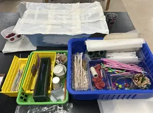 A photo showing a medium-sized green container with materials including popsicle sticks and paper cups, and a larger blue container with materials including chopsticks, yarn, string, duct tape, and pipe cleaners. 