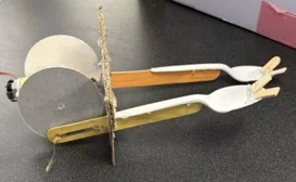 A photo showing a model of digging tool made up of cardboard, popsicle sticks, a plastic spoon, and a motor. The motor is in between two circular pieces of cardboard, a rectangular piece of cardboard is attached on the circumference of the circular pieces, a popsicle stick is attached on the surface of each circle at the side, and it passes through the rectangular piece of cardboard, then each popsicle stick has a plastic spoon attached to it. That head of each spoon has two small pieces of popsicle stick attached to it.