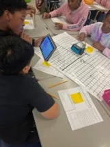 A photo showing a group of students sitting at desks, with one student holding the tablet to program an mBot2 robot while it is sitting on a large, printed coordinate plane worksheet.