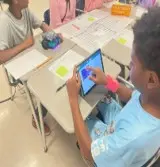 A photo showing a student holding a tablet and programming an mBot2 robot across from a student holding the mBot.