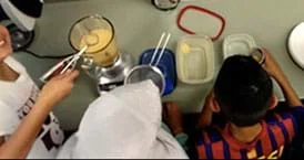 A photograph from above a classroom counter with a blender, mesh sieve and many containers shows three students crowding around the algal cell edible model setup.