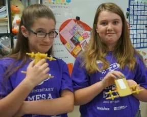 Two young people stand in a classroom, each smiling and holding in their hands four-wheeled vehicles made of dry pasta.