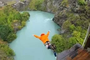 A person in an orange jacket, with a bungee cord attached to his ankles jumps off a platform headfirst into a gorge with a river at its base.