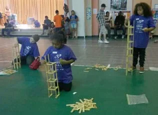 A photograph in a large room shows two girls and a boy individually stacking blocks on top of each other to make their own tall towers.