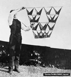 A black and white photograph shows a man using two hands to hold up and away from his body a 16-celled tetrahedron kite. The design includes opposing triangular vanes and was photographed outside against a white sheet.