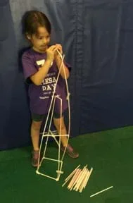 A photograph shows a young girl using plastic drinking straws and tape to build a tower structure that is almost as tall as she is.