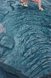 Photo shows a person standing on a wide swath of black rock that is textured with ripples and ridges.