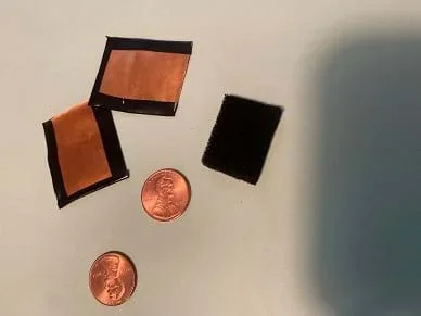Two one-inch square pieces of copper flashing with electrical tape around 3 sides, two pennies, and one piece of black conductive foam approximately the same size as the copper.