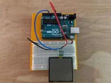 A square force-sensitive resistor is connected via a voltage divider circuit with a 10 k resistor built on a solderless breadboard and connected to an Arduino microcontroller.