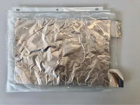 A photograph shows the stack of tin foil and wax paper together placed inside of a clear sheet protector so that the tin foil tails are sticking out of the sheet protector.