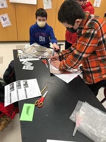 A photograph shows students around a table building a sensor. A student at the top of the photo sits in a chair and is grasping a piece of tin foil. A student is standing next to him watching. Another student to the right is using a marker to draw a line to cut out a tin foil piece for their sensor.