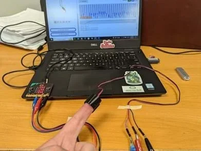 A photo showing a connected micro:bit and pulse sensor recording heart rate. A heartbeat waveform is generated on the screen in the background.