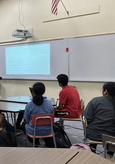 A photo showing three students sitting in chairs as they look at Python code displayed on a whiteboard.