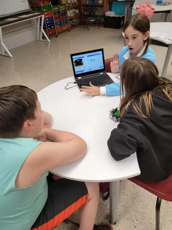 A photo showing three students working at a white round table in a classroom. One is sitting behind an open laptop and speaking. A second sits to the right, looking toward the laptop and a small robot on the table. The third sits in the foreground on the left, watching the screen, which displays a block-based programming interface.