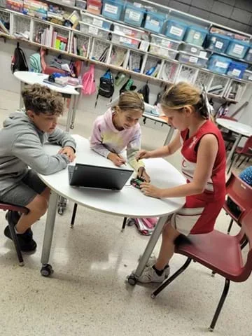 A photo showing three students clustered around a white circular table. They are collaborating on a STEM project involving an open laptop and a small, wheeled Cutebot robot.