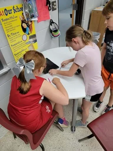 A photo showing three students working together at a small round table in a classroom. One student sits in a chair using a laptop while writing with a pencil, while another leans over the table assisting with the computer. A third student stands nearby observing. The scene shows collaborative problem-solving in a classroom environment with posters and school materials in the background.