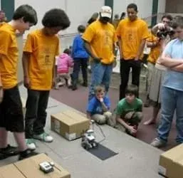 Students and teachers gather around a LEGO robot on a classroom floor along with various cardboard boxes.