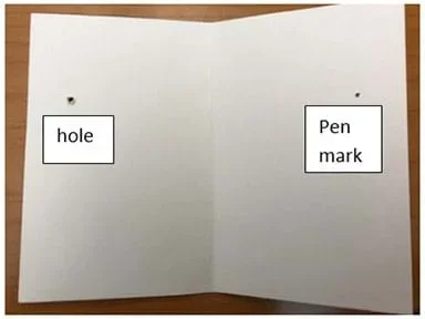 Photograph of an open piece of blank cardstock showing the inside where a hole has been poked through the left side with a pen point and a pen mark has been marked through the hole on the right side.