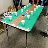 A photo showing a small rectangular box being pulled by a string along a table covered with green paper, with a strip of brown paper down the middle, surrounded by 12 paper cups.