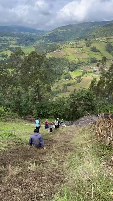 A photo of the same grassy hillside viewed from the top, where people are lined up along a now worn down and muddy path in the grass. A vast agricultural and forested valley near the Yambiro community of Otavalo, Ecuador is in view.