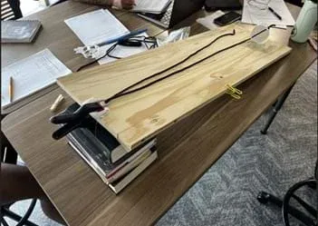 A photo of a student project demonstrating an inclined plane or a similar physics principle using a wooden plank, a stack of books, and a simple pulley system with a string and a white cup.