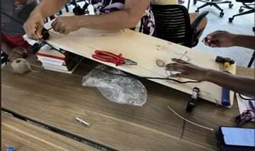 A photo showing students working to set up an inclined plane using a wooden plank propped on books, with multiple hands adjusting the black string and a white plastic cup (the load). Various tools and materials, including pliers, a hammer, and twine, are scattered nearby as they prepare or test the setup.