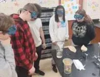 A photo of five students standing around a lab desk. There are three beakers, two of which have funnels in them. Two pieces of filter paper and a strip of toilet paper are also on the desk.