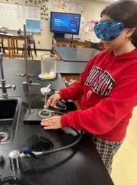A photo of a student standing in front of a Bunsen burner and ring stand. A beaker with a yellow leaf in it is on top of the wire mesh on top of the ring clamp clamped to the ring stand.
