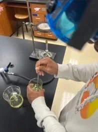 A photo of a student stirring a beaker with a yellow fluid in it. A Bunsen burner and ring stand are set up nearby.