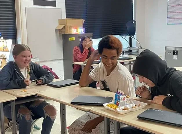 A photo showing three students sitting at a table in a classroom, two boys and one girl. One of the boys is attempting to suck up a Goldfish cracker with a straw while the other two laugh.