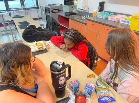 A photo showing three girls sitting at a table in a classroom, two with their back to the camera and one looking down at the desk. One of the girls is attempting to move Goldfish crackers with her straw while the other two are watching.