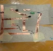 A photo of an OR gate built on a piece of cardboard from a cereal box cut to the size of a notecard. The gate is built using copper tape, three resistors, two transistors and an LED. There is also masking tape used to separate layers of the copper tape to prevent short circuits. There are two places in the circuit where gaps are left between pieces of copper tape to create the switches in their open states.
