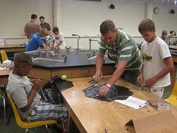 Image of a teacher standing at a counter in a science lab with two young students helping them create parachutes out of black plastic bags and string. The lab table is has supplies on it such as clipboards, paper, pens, tape and safety goggles.