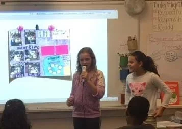 Two female fifth graders stand at the front of the classroom. The one on the left is speaking into a microphone. They have their poster presentation illuminated and hung on the board. The poster has pictures and writing, and is visually appealing. Other students sit and listen.