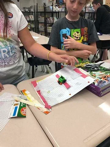 Two students test elements of friction using a green toy car, and an inclined ramp set up on a textbook.