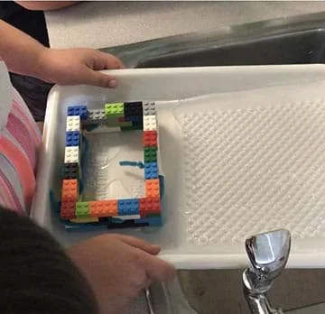 The photo shows a paint tray holding a house made of plastic bricks. Water is flowing into the tray. The plastic house has pipe cleaners and tape protruding from two windows.