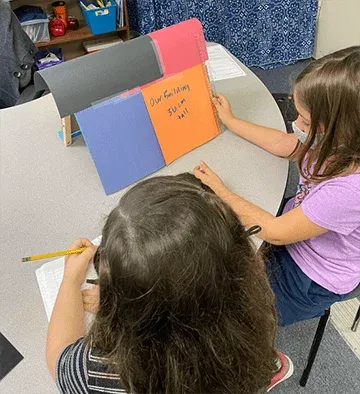 One student is measuring their construction paper building with a ruler, which reads thirty-six centimeters. Another student is recording their data in a notebook.