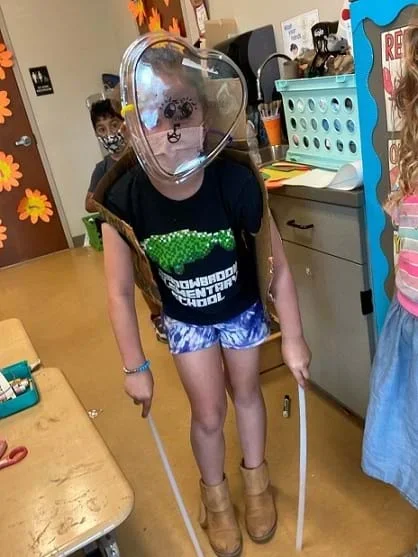 A child shows off an example of an animal costume. There is a shell made out of cardboard on their back and a clear plastic mask with an animal face on their head. The child is holding two poles pointed down at the floor to imitate long legs.