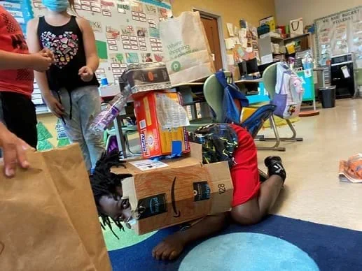 A student shows off an example of an animal costume. The child is on the ground on their elbows and knees. There is a large, shell-like costume on their back made out of cardboard with a plastic bottle attached to the front.