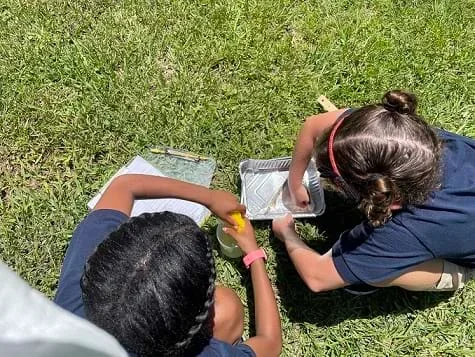 A photograph shows one student squeezing water and oil into a beaker using a sponge. Another student next to her is using a cotton ball to absorb the water and oil.