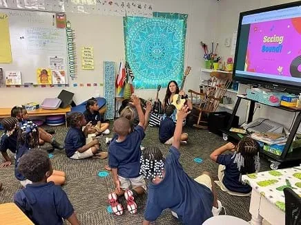 A teacher shows an elementary school class a guitar and demonstrations sound vibrations.