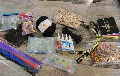 A photograph of craft materials, including pipe cleaners, paper clips, fabric mesh, beads, yarn, glue, bungee cords, rubber bands, and felt sheets.