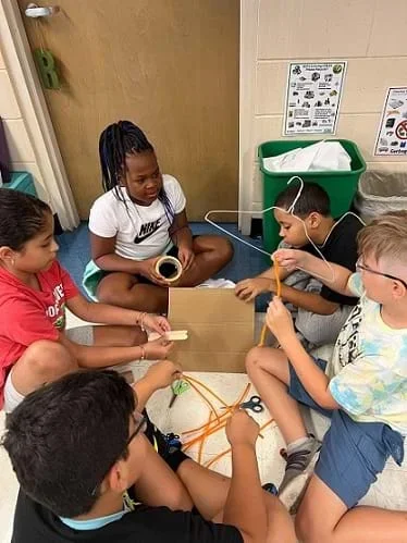 A group of five students sit in a circle, each holding various items for their design, including a hanger, wood blocks, and cardboard.