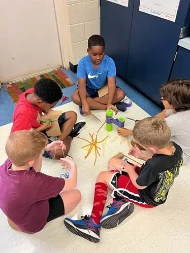 A group of five students sit in a circle and work on their design using teacher-provided materials.